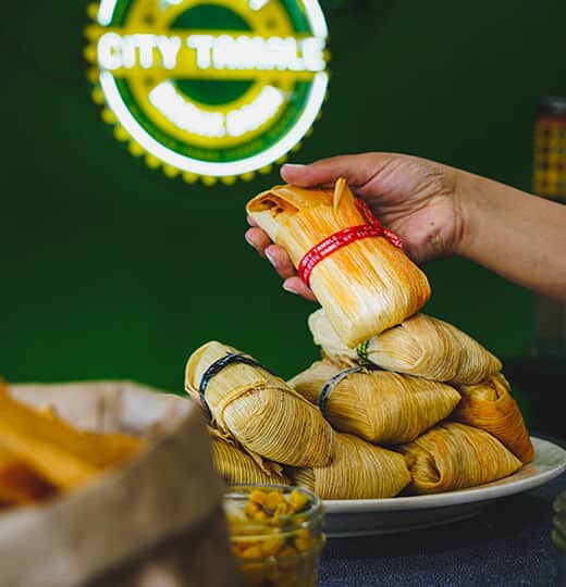 A hand reaching for a pile of wrapped tamales in front of the City Tamale sign